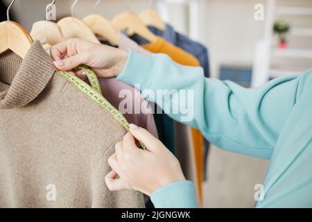 Close-up of unrecognizable female dressmaker in sweatshirt standing at clothes rack and using measuring tape to check measurements of sweater Stock Photo