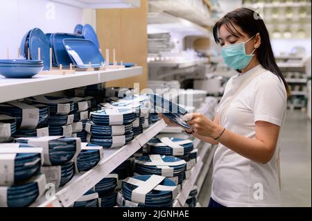 Asian woman wearing a protective mask chooses plates in tableware store Stock Photo