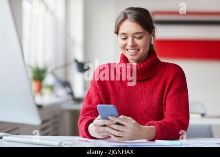 Smiling beautiful young woman in wireless earphones sitting at desk with computer and testing UX design on smartphone Stock Photo