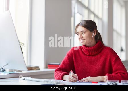 Smiling attractive young woman in red knitted sweater and earbuds sitting at table and using computer while working on web design project Stock Photo