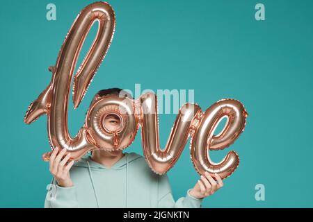 Portrait of playful young woman in hoodie standing against teal background and looking through letter O of foil balloon in shape of love word Stock Photo