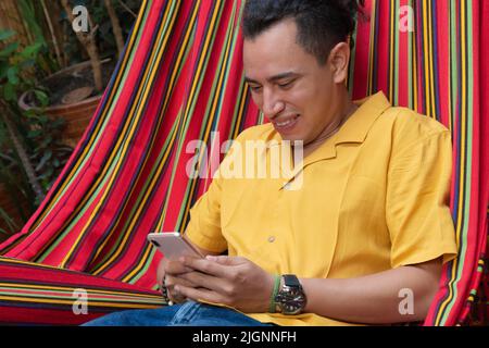 Latin Man resting in a hammock at home and uses a smartphone. Stock Photo