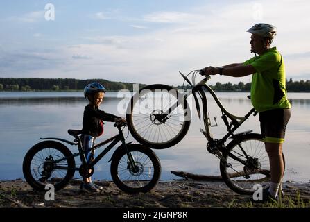 Silhouettes of an adult man and a boy with two bicycles in helmets near the lake at sunset. cycling, healthy lifestyle. Be like dad. Family pastime, d Stock Photo