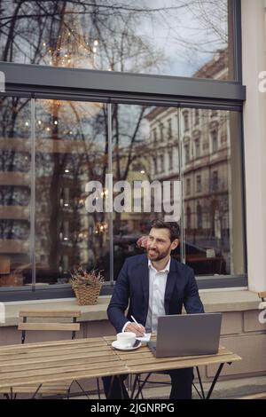 Attractive elegant man taking notes while working at laptop in cafe outside. Coffee break. Stock Photo