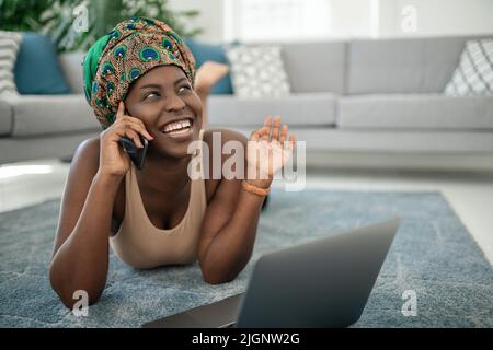 Beautiful African woman at home, smiling and laughing, using mobile smart phone, wearing traditional headscarf, surprised and happy Stock Photo