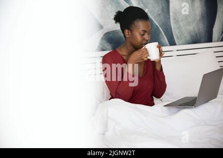 Young beautiful African woman sitting in bed wearing pajamas, sipping on cup of coffee and watching movies on laptop Stock Photo