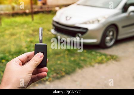 Man's hand pushes a button on the car remote control against parked blurred car. Vehicle key in male hand outsides. Unlocking vehicle by remote key si Stock Photo
