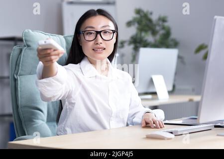 Heat in the workplace. A young Asian business woman in glasses, a white shirt sits in the office at the desk, turns on the air conditioner with the remote control, needs fresh air. Stock Photo