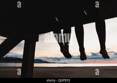Silhouette feet of couple sitting on the pier at sunset beach. Stock Photo