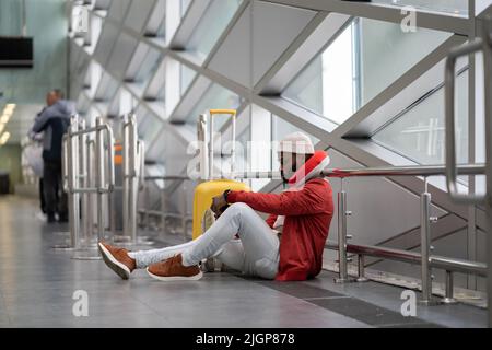 Concentrated young black man sitting on floor airport near window using mobile phone waiting flight. Stock Photo