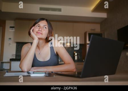 asian female freelancer working on laptop in living room. Focused lady studying distantly and making notes in copybook, listening to lecture. technolo Stock Photo