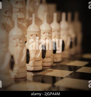 Blurred image.Carved vintage wooden chess pieces like vikings stand on old chessboard in starting position before the battle. Strategic logic games Stock Photo