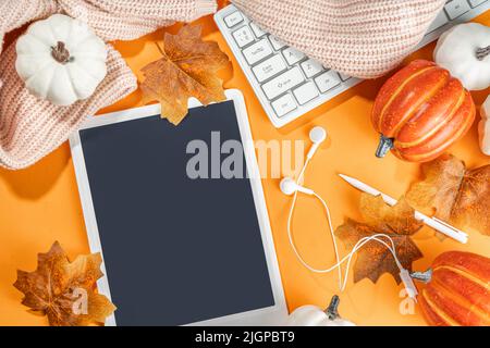Cozy fall background with tablet, ear-pods, white, orange pumpkins, autumn leaves decor on high-colored orange background. Autumn still life compositi Stock Photo