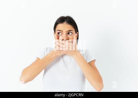 Worried young brunette woman covers her mouth with her hands timidly looking around, afraid to speak Stock Photo