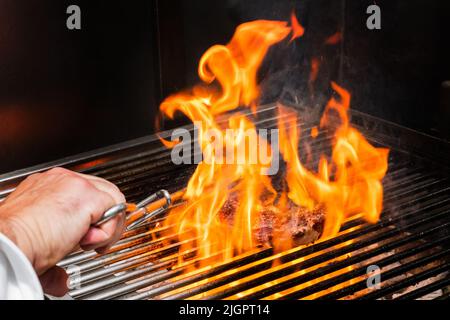 Veal cutlet on the grill with flames of fire and hand with tongs Stock Photo
