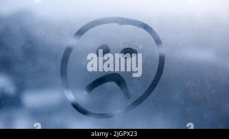 Hand drawn sad emoji on foggy glass window background. Unhappy face sign close-up. Selective focus on photo. Depression and sadness concept. Stock Photo