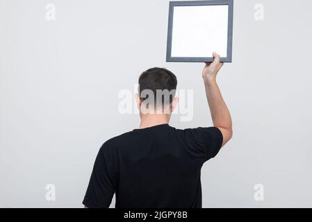 Back view of man holding blank board with black frame over head on white background. Copy space. Back man wearing black t-shirt. Stock Photo
