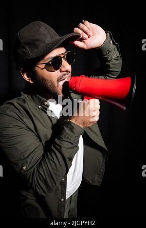indian hip hop singer in sunglasses and cap screaming in megaphone on black Stock Photo