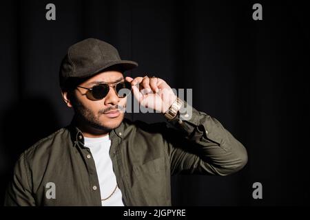 eastern hip hop performer in cap adjusting sunglasses on black Stock Photo