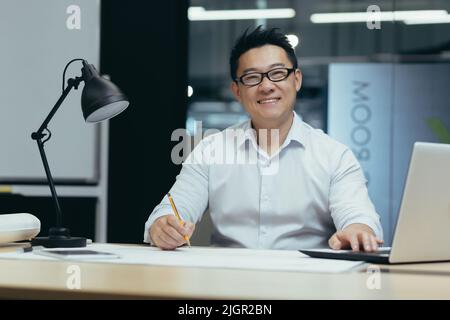 Portrait of Asian designer, creative work, man looking at camera and smiling, working in architecture office on plan drawing Stock Photo