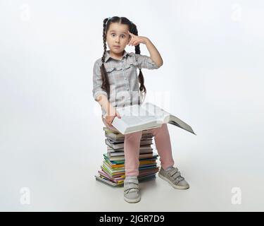 charming little girl with braided pigtails sits on a large stack of books and holds her finger near her head in surprise. Stock Photo