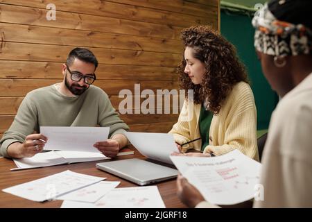 Three young intercultural economists looking through financial papers and analyzing charts, graphs and diagrams at meeting in office Stock Photo