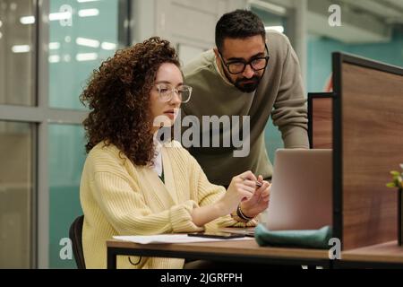 Two young colleagues in eyeglasses and casualwear looking through data on laptop screen during working meeting in office Stock Photo