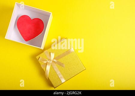 Open gift box in gold color with ribbon and bow with heart inside on yellow background Stock Photo