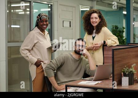 Three young successful intercultural colleagues with mobile gadgets looking at camera while working over new business project Stock Photo