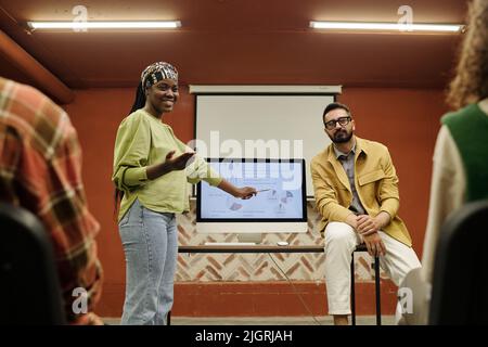 Happy young African American speaker pointing at computer screen with diagrams and other financial data while explaining it to colleagues Stock Photo