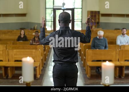 Rear view of young African American priest in black shirt and pants blessing parishioners sitting on benches during church service Stock Photo