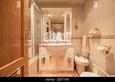 Small bathroom of home with white shower stall, wall mirror and porcelain sink on white wooden cabinet and beige tiles on the wall Stock Photo