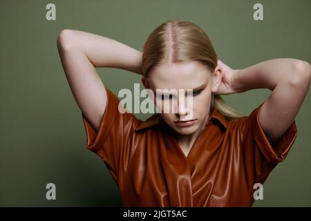 Fashionable stylish woman touching hair posing isolated in brown palette leather dress trench coat isolated on over olive green wall background. Cool Stock Photo