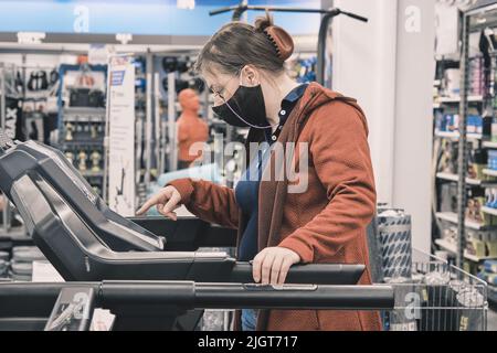 Woman wearing glasses and a mask against virus buys a large treadmill in a shop Stock Photo