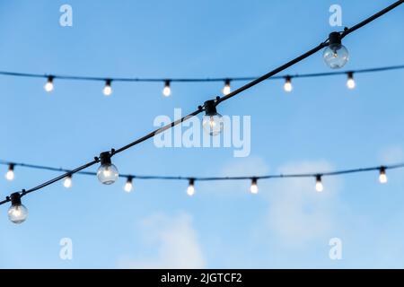 Street garlands of light bulbs hanging on black wires are under blue evening sky Stock Photo