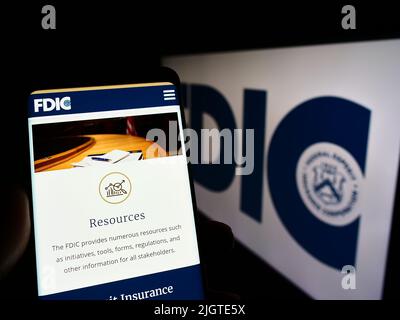 Person holding cellphone with webpage of US Federal Deposit Insurance Corporation (FDIC) on screen with logo. Focus on center of phone display. Stock Photo