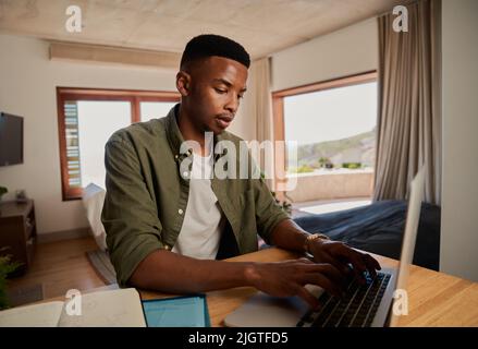 Focused young adult African-American black male typing on laptop while working remotely. Remote work and Isolating in modern apartment. Digital Nomad Stock Photo