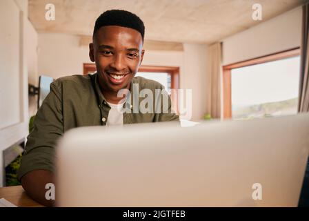 Close up portrait of a happy Young adult black African-American male smiling while using his laptop. Remote working from his modern apartment Stock Photo