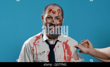 Brain-eating zombie with creepy smirk getting vaccine shot on arm. Horror looking dead walking corpse with bloody and deep wounds getting vaccinated while smiling at camera on blue background. Stock Photo