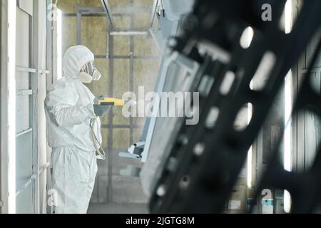 Industrial worker in protective workwear using spray gun to paint metal details at factory Stock Photo