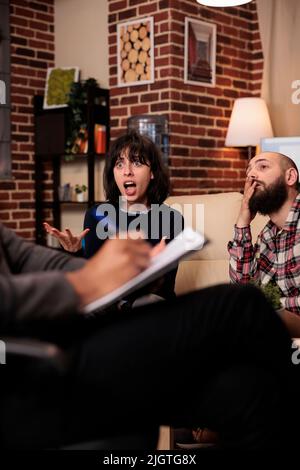 Male psychologist taking notes on paper at couple therapy with angry partners, talking about relationship issues. Doing psychoanalysis with married people, giving counseling advice. Stock Photo
