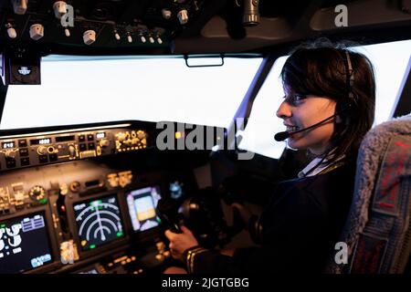 Aircrew member flying plane from cockpit with dashboard command and control panel, using steering wheel and control panel for windscreen navigation. Woman using lever to fly aircraft. Stock Photo