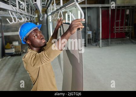 African young worker in hardhat hanging sheets on rack during his work at industrial factory Stock Photo