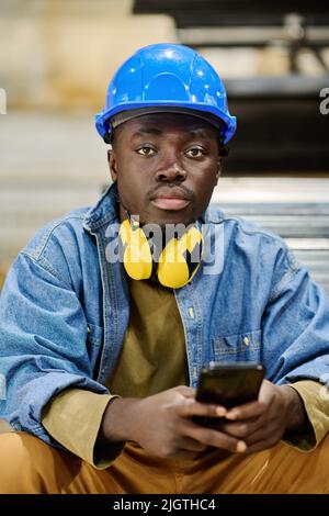 Portrait of African young male worker in protective hardhat looking at camera while playing on his smartphone during break Stock Photo