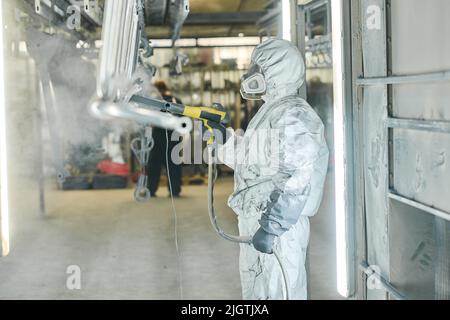 Coveralls worker with spray gun painting steel parts in painting chamber Stock Photo