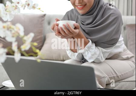Beautiful elegant young Asian Muslim woman holding a hot coffee cup while looking at her work on laptop screen. cropped image Stock Photo