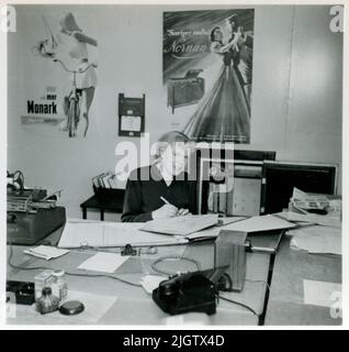 The office at Monarch's bicycle workshop. A woman sits at a desk and writes in a cover. On the wall behind her is a poster with advertising for Monarch's bikes and one for Monark's radio furniture "Swedish Radio Nornan". Stock Photo