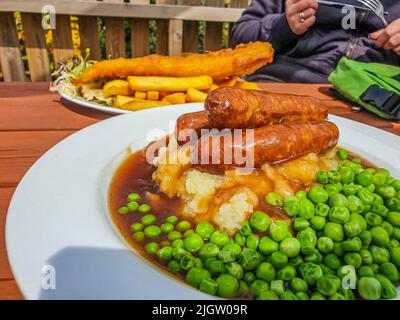 Bangers (Sausages) and Mash with Onion Gravy and peas. Fish and chips in the background at Devonshire Pub, UK Stock Photo