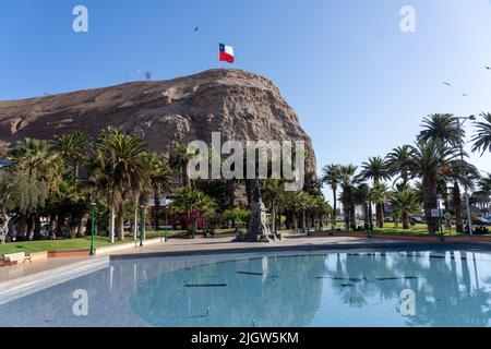 A giant Chilean flag flies on top of the El Morro hill overlooking a park in Arica, Chile.  A statue of Benjamin Vicuna Mackenna and a pool are in the Stock Photo