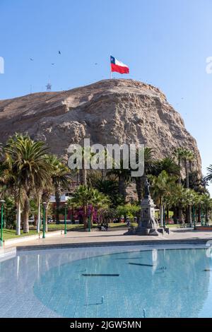 A giant Chilean flag flies on top of the El Morro hill overlooking a park in Arica, Chile.  A statue of Benjamin Vicuna Mackenna and a pool are in the Stock Photo